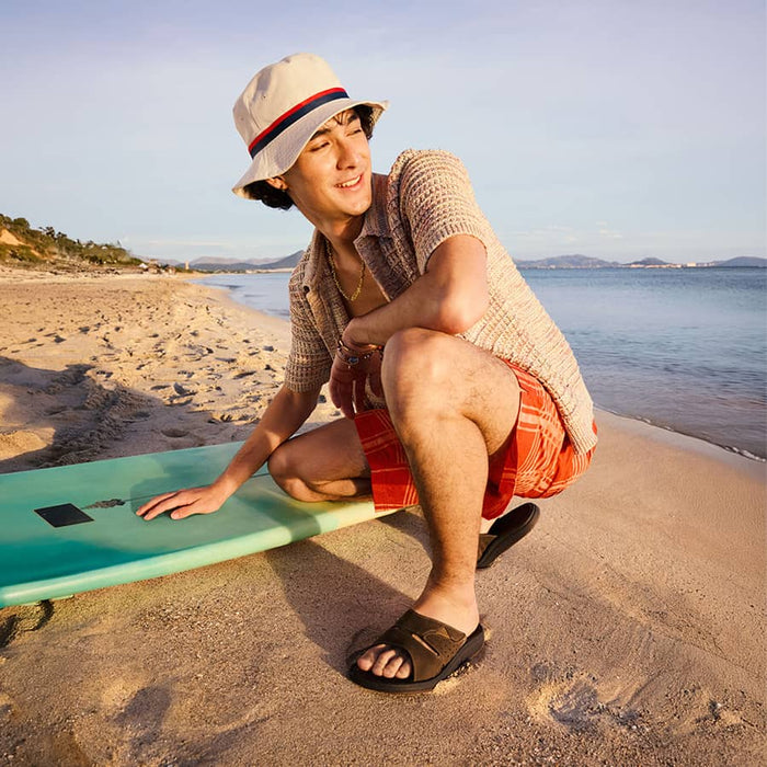 A young man in a beige bucket hat, red shorts and Clarks Brinkley Slide Sandals sits on the beach next to a turquoise surfboard, smiling while resting his hand on the board.